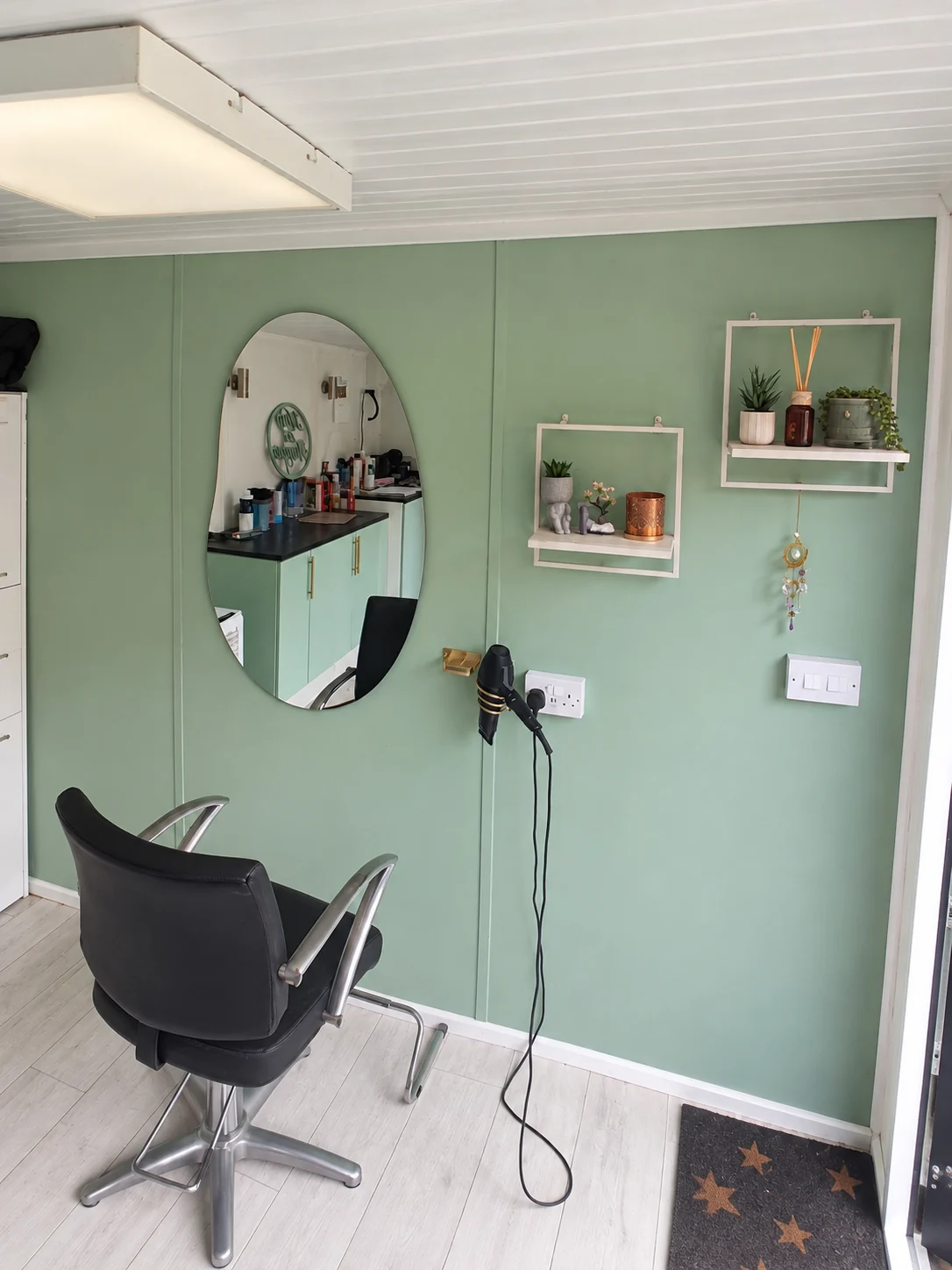 Inside my garden salon, with a sage green wall, oval mirror and black styling chair